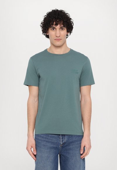 Camiseta de algodón color aguamarina con cuello redondo y mangas cortas. Presenta un logotipo sutil en el pecho. Combinada con jeans de mezclilla azul.