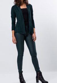 Blazer texturizado de color verde oscuro, con mangas largas, apertura frontal y cierre con botones, combinado con pantalones brillantes de color verde oscuro y botas negras hasta el tobillo.
