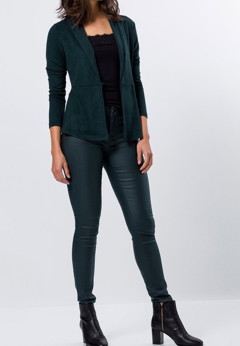 Blazer texturizado de color verde oscuro, con mangas largas, apertura frontal y cierre con botones, combinado con pantalones brillantes de color verde oscuro y botas negras hasta el tobillo.