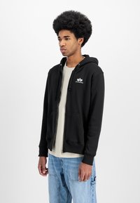 Sudadera negra con cremallera y logo frontal, con bolsillos tipo canguro y puños de canalé. Llevada sobre una camiseta de color claro y combinada con vaqueros azules.