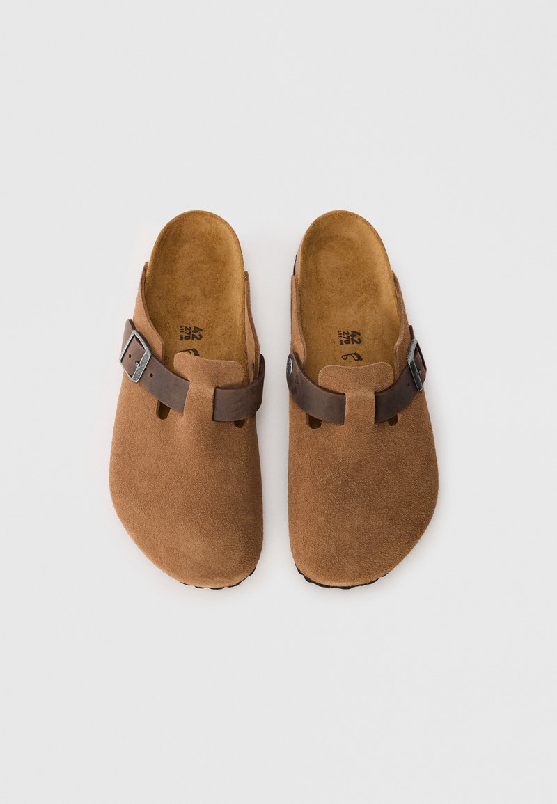 Sabots en daim marron avec un bout arrondi, une sangle en cuir marron et une boucle en métal argenté, dotés d'une semelle intérieure douce et d'une semelle extérieure texturée.