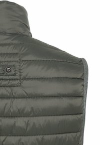 Gros plan de la partie supérieure du dos d'un gilet matelassé vert foncé avec des coutures horizontales et un petit détail boucle en tissu.