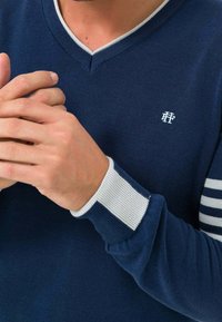 Marineblauer V-Ausschnitt-Pullover mit gerippten Bündchen. Die Bündchen haben weiße Streifen, und das Logo auf der Brust ist weiß. Glatte, gestrickte Textur.