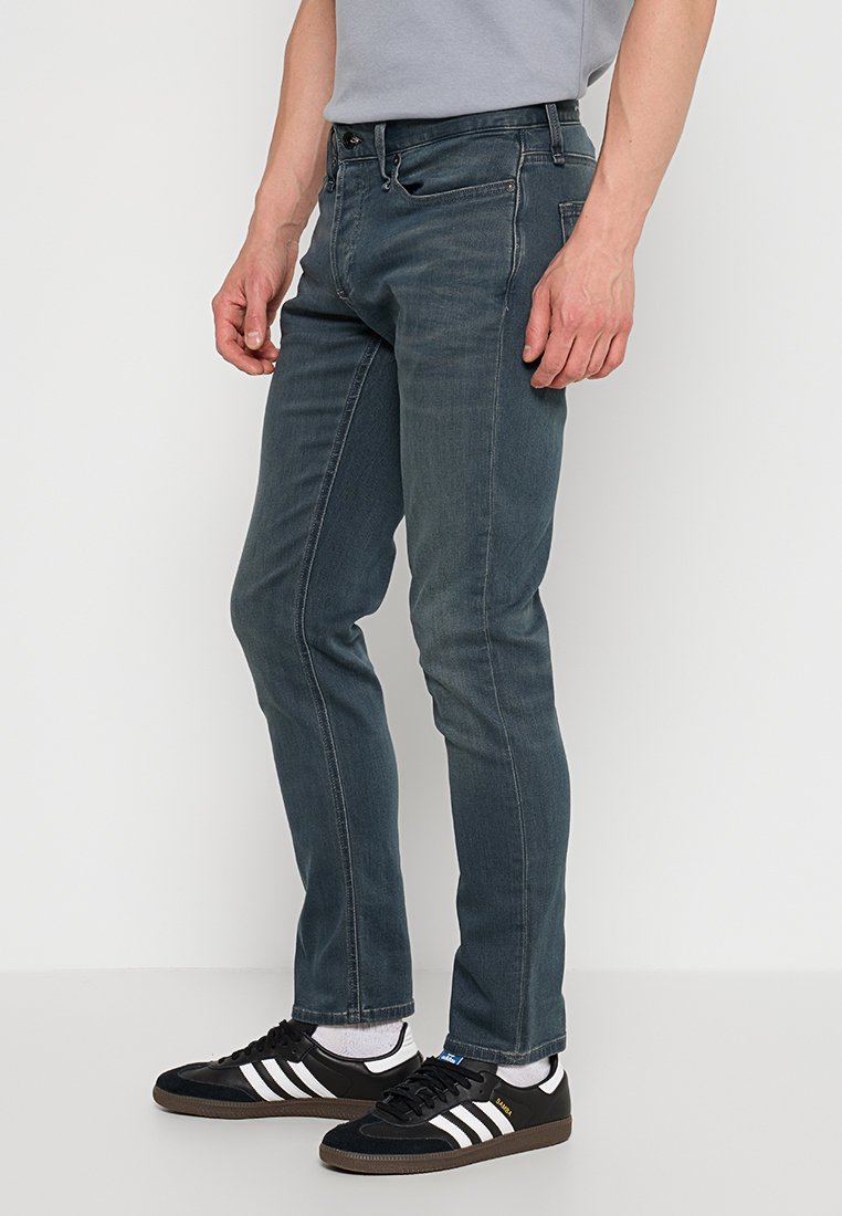 Man in slim-fit blauwe jeans en zwart-witte sneakers, staande tegen een plain witte achtergrond.