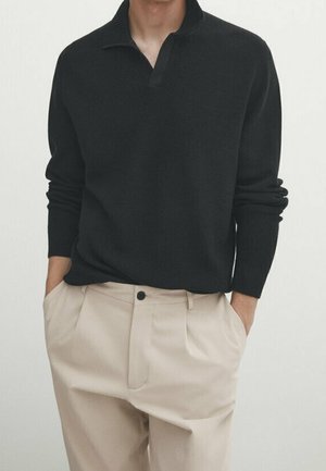 Man wearing a black long-sleeve knit sweater with a collar and beige high-waisted pleated trousers, hands in pockets.