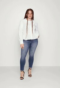 Mulher com tranças longas, vestindo uma blusa branca com um grande colarinho, calças de ganga justas azuis e sandálias de salto alto pretas, de pé contra um fundo simples.