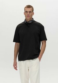 Sort kortærmet t-shirt med en afslappet pasform, der har en rund halsudskæring. Sat sammen med cremefarvede bukser og et sort bandana om halsen.