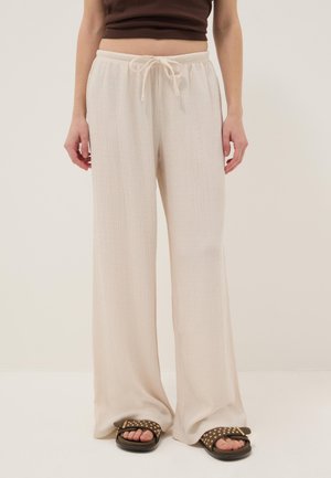 Vrouw draagt losse crèmekleurige broek met trekkoord en bruine sandalen met studs, staand tegen een neutrale achtergrond.