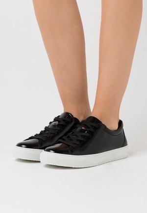 Zapatillas de charol negro con suelas de goma blanca, que presentan puntera redonda, cordones texturizados y acentos de logo en los laterales.