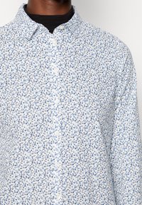 Camicia di cotone bianca con un motivo floreale blu, dotata di colletto classico e chiusura frontale con bottoni. Tessuto morbido e vestibilità regolare.