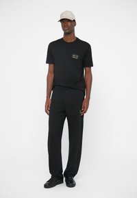 Juodas medvilninis marškinėliai su mažu logotipu, derinami su juodomis džersio kelnėmis ir šviesiai rudais kepurėle. Batai juodi, su paprastu dizainu.