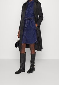 Person som bär en jeansklänning med midjebälte, en lång svart trenchcoat, knälånga svarta läderstövlar och håller en liten svart handväska.