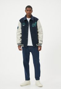 Giacca varsity blu navy con maniche color crema, decorata con la lettera "A" ricamata in verde, strisce ai polsini e loghi laterali. Abbinata a jeans blu.