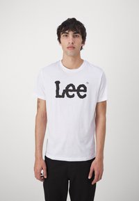 Hvit bomull t-skjorte med en fremtredende svart grafisk logo "Lee" i en fet, stilisert skrift. Korte ermer og rund hals.