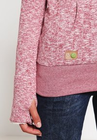 Punainen ja valkoinen täplikkäinä huppari, joka on valmistettu tekstuurikankaasta. Hupussa on ribattu helma ja logo-patch etuosassa.