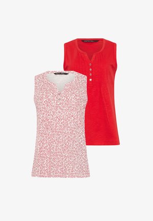 Kvinde med krøllet hår iført ærmeløs lyserød blomsterbluse, blå jeans og brune sandaler, stående med den ene hånd i lommen.