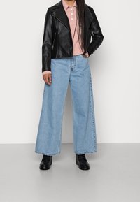 Svart lädermotorcykeljacka, rosa stickad knapplöst, vida ljusblå jeans och svarta ankboots, framför en enkel bakgrund.