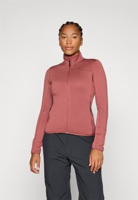 En kvinne poserer iført en burgunder zip-up jakke og sorte bukser, med håret stylet i fletter, mot en enkel hvit bakgrunn.