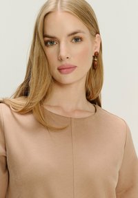 Blonde Frau mit grünen Augen, die ein beiges, strukturiertes Oberteil und braune Ohrringe trägt, vor einem schlichten hellen Hintergrund.