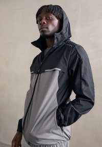 Veste de sport pour homme présentant un design en blocs de couleur noir et gris, fermeture éclair à l'avant, capuche et accents réfléchissants subtils. Fabriquée en tissu léger.