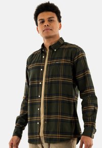 Groene en gele geruite button-up shirt met een borstzak en witte knopen, gelaagd over een beige shirt. Het heeft lange mouwen en een kraag.