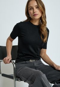 Camiseta de punto negra de manga corta con cuello de canalé, combinada con jeans grises sueltos. Modelo sentado en una silla de cromado.
