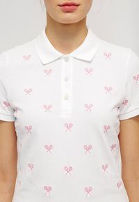 Vit polo skjorta gjord av texturerad tyg, med ett mönster av små rosa tennisrackets, två knappar och en klassisk krage.