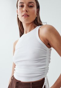 White, ribbed tank top with adjustable side ties and a fitted design. Finished with a scooped neckline and soft texture.