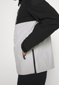 Černá a světle šedá bunda se zipovou kapsou, vysokým límcem, nastavitelnými rukávy a strukturovanou tkaninou na rukávech.