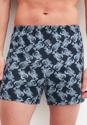Mann trägt marineblaue Boxershorts mit einem wiederholenden hellblauen Hummermuster, stehend vor einem schlichten Hintergrund.