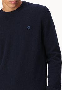Granatowy sweter z okrągłym dekoltem wykonany z miękkiego materiału, z długimi rękawami i małym, tonowym logo na klatce piersiowej. Klasyczny, minimalistyczny design.