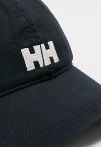 Donkerblauwe pet van katoen met een gebogen rand. Voorzien van een witte geborduurde "HH" logo aan de voorkant en zichtbare stikdetails.