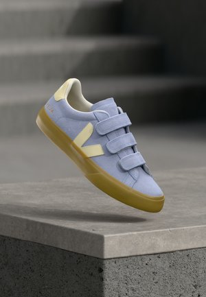 Light blue suede sneaker with beige accents and three velcro straps, floating above a concrete ledge against blurred gray stairs.