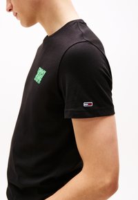 Tricou din bumbac negru cu un logo verde pe piept și un mic eticheta colorată pe mânecă. Design cu guler rotund și mâneci scurte.