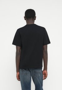 Μαύρο κοντομάνικο μπλουζάκι με στρογγυλή λαιμόκοψη, κατασκευασμένο από απαλό βαμβάκι, με ελάχιστες ραφές και χαλαρή γραμμή. Μπλε τζιν παντελόνι ορατό από κάτω.