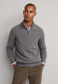 Maglione grigio lavorato a maglia con colletto a zip quarti, polsini e orlo a coste. Indossato sopra una camicia a righe bianco e blu chiaro.