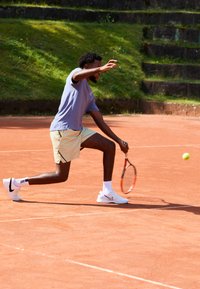 Tennis spiller i lyseblå skjorte og stribede shorts lunges på grusbanen og svinger sin ketsjer mod en gul bold, iført hvide Nike sko.
