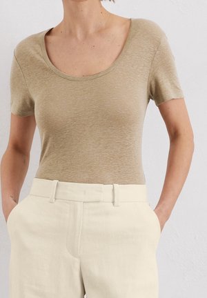 Femme portant un haut beige à manches courtes rentré dans un pantalon taille haute blanc cassé, les mains dans les poches, sur un fond uni.