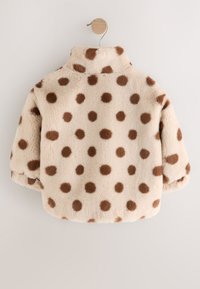 Veste en fausse fourrure beige avec des pois marron, un col haut et une texture douce. Suspendue sur un crochet simple.