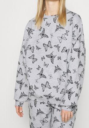 Persona vestida con sudadera y pantalones a juego de color gris claro, cubiertos con patrones de mariposas negras, de pie contra un fondo liso.