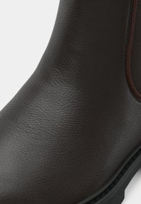 Gros plan sur une botte en cuir brun foncé présentant un dessus texturé, un panneau latéral élastique et une partie de la semelle noire sur fond blanc.