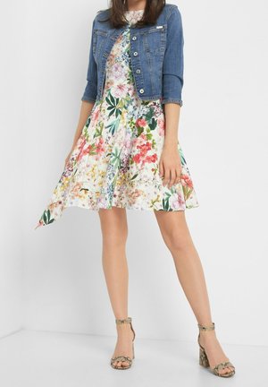 Vestido floral con mangas cortas, con patrones vibrantes en rosa, verde y azul; combinado con una chaqueta vaquera corta y sandalias de tacón.