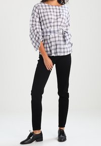 Donna che indossa una blusa a quadri bianca e viola con cintura e maniche a tre quarti, pantaloni neri slim e scarpe nere con lacci.