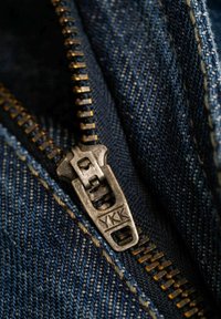 Primer plano de una cremallera de metal en denim azul oscuro, con un tirador YKK, costuras visibles y detalle de tejido texturizado.