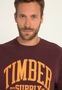 Bordowy sweatshirt z wyraźnym żółtym napisem "TIMBER SUPPLY" oraz datą założenia. Miękka tkanina, klasyczny krój z okrągłym dekoltem.