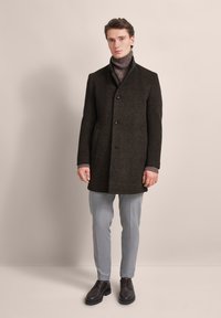 Dark brown wool coat with a high collar, front buttons, and side pockets, layered over a textured sweater and paired with light gray trousers.