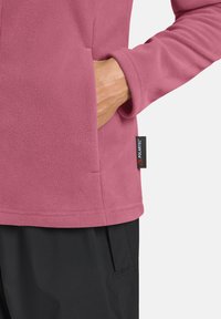 Giacca in fleece di un delicato rosa con colletto arrotondato, tasche laterali e un'etichetta nera POLARTEC sull'orlo; presenta una texture liscia.