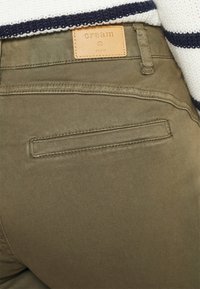 Calças verde-oliva feitas de tecido suave, com um retalho de couro rotulado como "creme" e um bolso traseiro com costura cuidada.