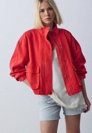 Junge Frau mit blonden Haaren, die eine knallrote übergroße Jacke, ein weißes Spitzenoberteil und helle Jeansshorts vor schlichtem Hintergrund trägt.