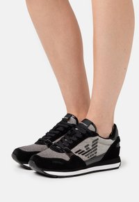 Jalka, jotka käyttävät mustia ja hopeita matalalattiaisia tennareita logo-suunnittelulla, seisovat valkoista taustaa vasten.